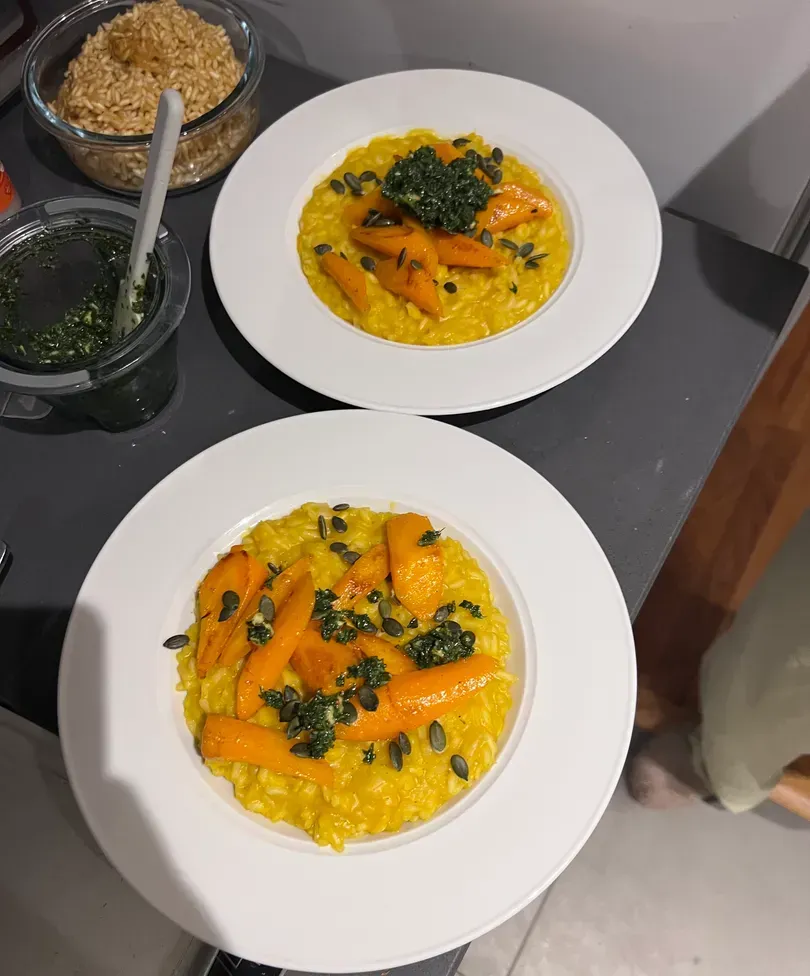 Two bowls of the risotto from above, with jars of carrot top pesto and pumpkin seeds beside them