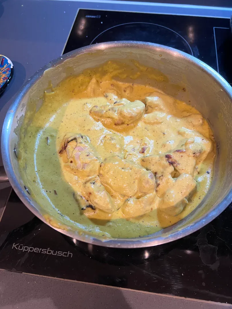 Chicken pieces in a pale yellow saffron and almond cream gravy in a steel pan on the hob
