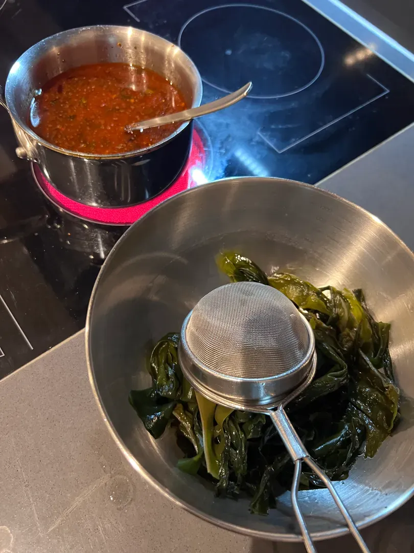 Kombu being lifted out of its soaking water with a sieve, chilli oil on the hob behind